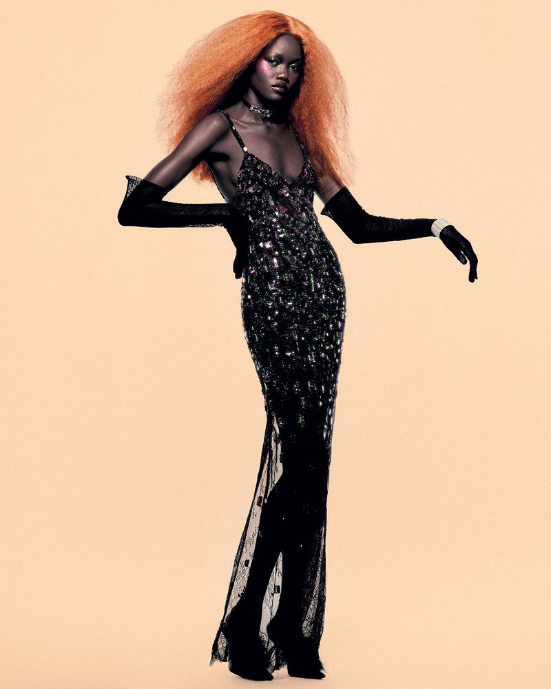 Model with vibrant orange hair wearing a sequined black slip dress and long black gloves against a pale backdrop
