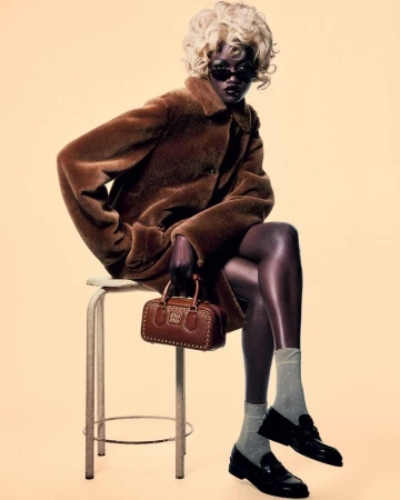 Model wearing a brown coat and sunglasses seated on a stool holding a handbag against a seamless background.
