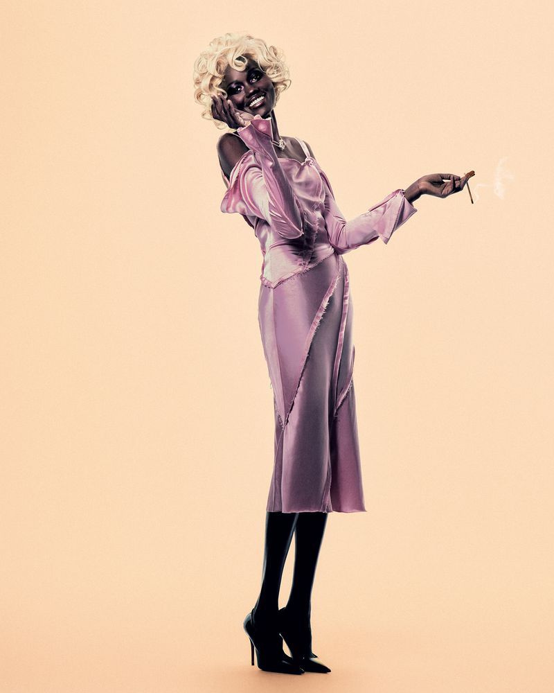 Model wearing a pink satin dress and blonde curly wig holding a cigarette against a plain warm background