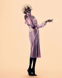 Model wearing a pink satin dress and blonde curly wig holding a cigarette against a plain warm background