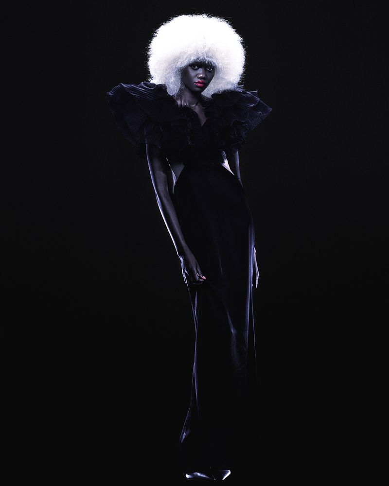 Model with large white afro hairstyle wearing a dark ruffle dress against a black background for AURUM project.