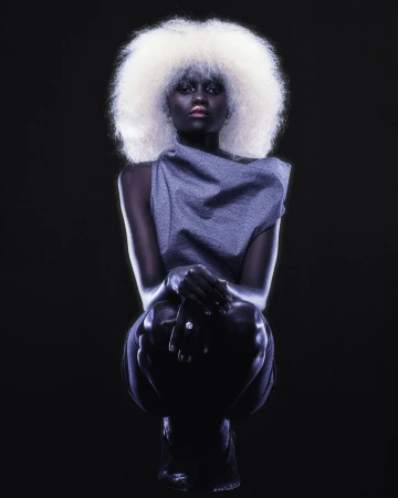 Model Caren Jepkemei with voluminous white afro hair wearing a grey top against a black background for the AURUM project