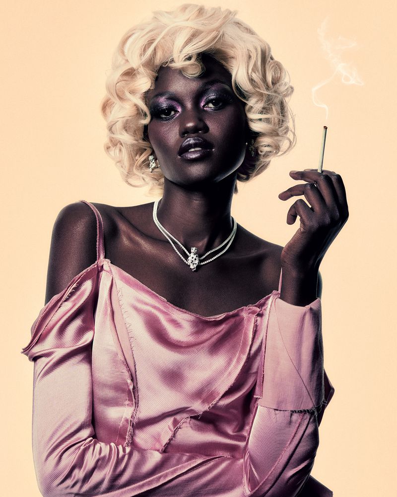 Black woman with blonde curly hair wearing a pink satin top and holding a lit cigarette against a neutral backdrop