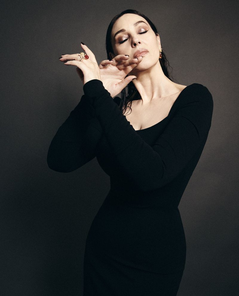 Monica Bellucci in a black long-sleeve dress with her hands posed gracefully near her face against a dark studio background