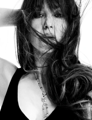 Portrait of Monica Bellucci with tousled hair wearing a black top and pendant necklace against a white background