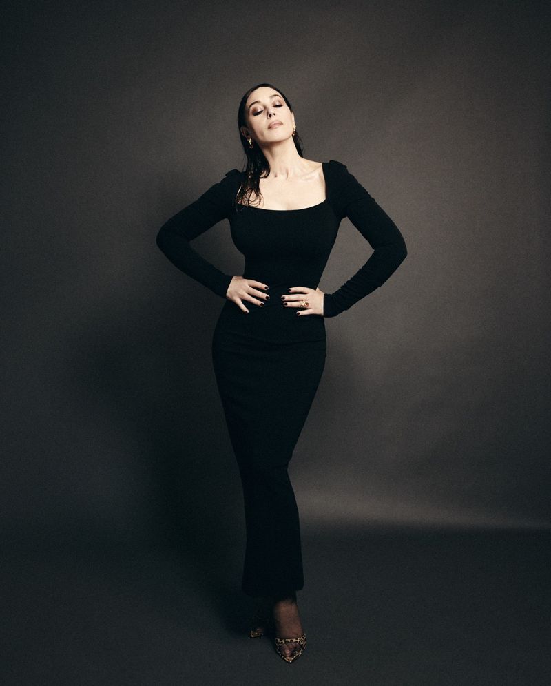 Monica Bellucci posing in a long black dress with hands on hips against a dark studio backdrop for Harper's Bazaar Espana.