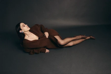 Monica Bellucci lying on a dark seamless studio backdrop wearing a brown knitted cardigan and heels