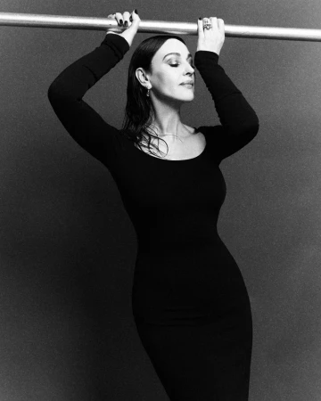 Monica Bellucci in a black dress holding onto a horizontal metal bar against a textured grey studio background.