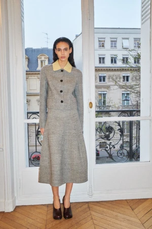 Model standing indoors in a grey two-piece skirt suit with a yellow collar in front of a window overlooking a city street.
