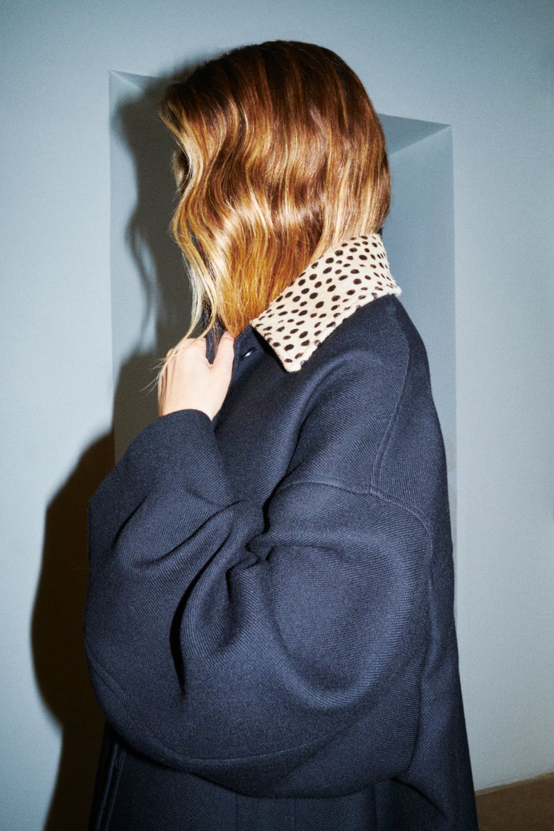 Model with highlighted hair wearing a dark coat with a dotted collar against a plain wall