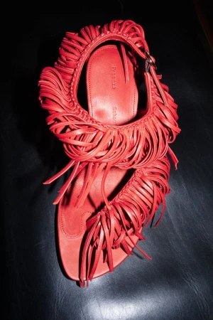 Close-up overhead shot of a vibrant red fringed sandal resting on a dark textured surface.