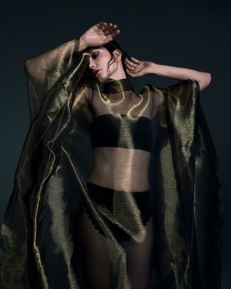 Actor Sylvia Hoeks posing in a sheer metallic gold cape and dark undergarments against a dark background