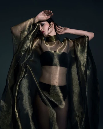 Actor Sylvia Hoeks posing in a sheer metallic gold cape and dark undergarments against a dark background
