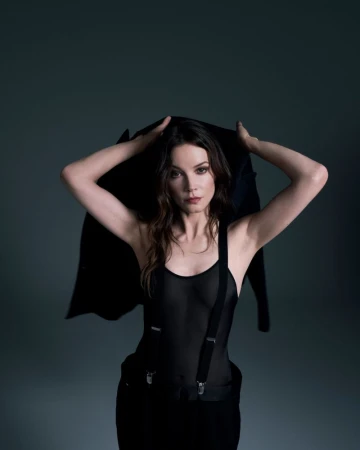 Sylvia Hoeks holding a black jacket behind her shoulders wearing a black top with suspenders against a dark studio background