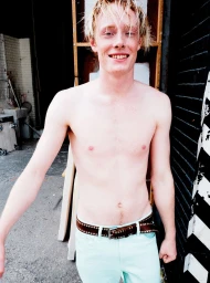 Shirtless young man smiling wearing light blue pants and a studded brown leather belt against an industrial background