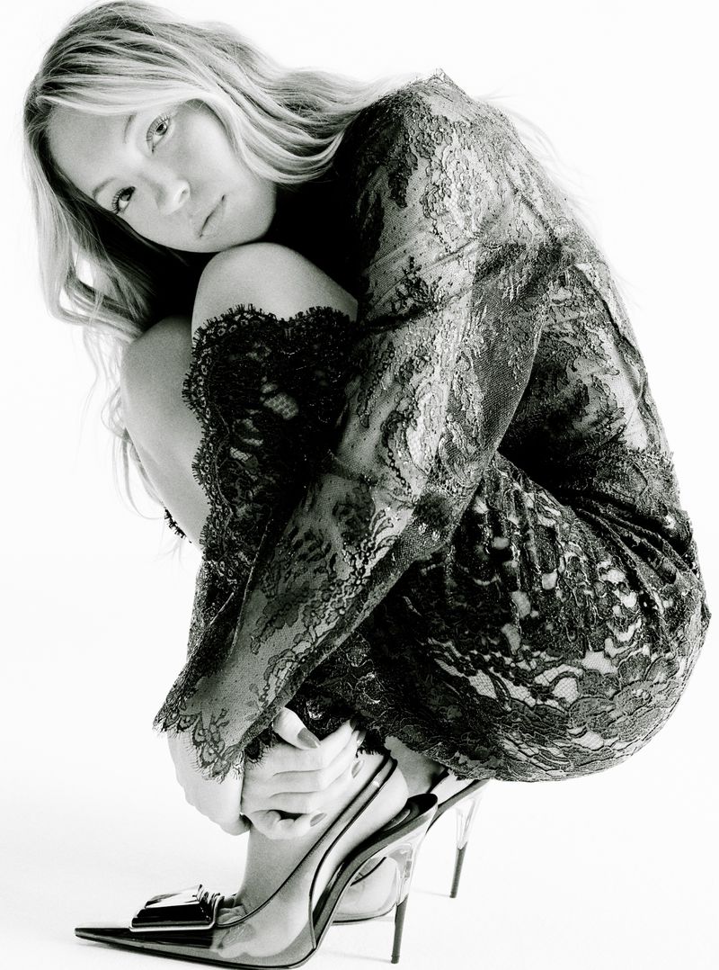 Model Kate Moss crouching while wearing a black lace long sleeve dress and pointed heels against a white background.