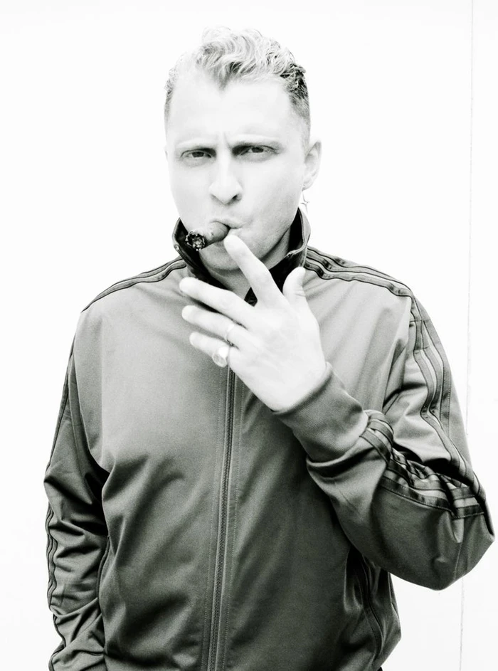 Monochrome portrait of a man wearing a zip-up track jacket with stripes on the sleeve, holding a cigar to his mouth.