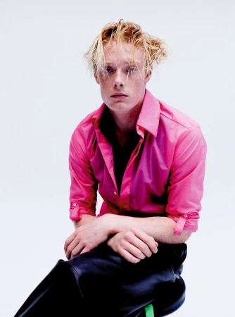 Young man with blonde hair and pink shirt sitting against a plain white background for The Perfect Magazine editorial.