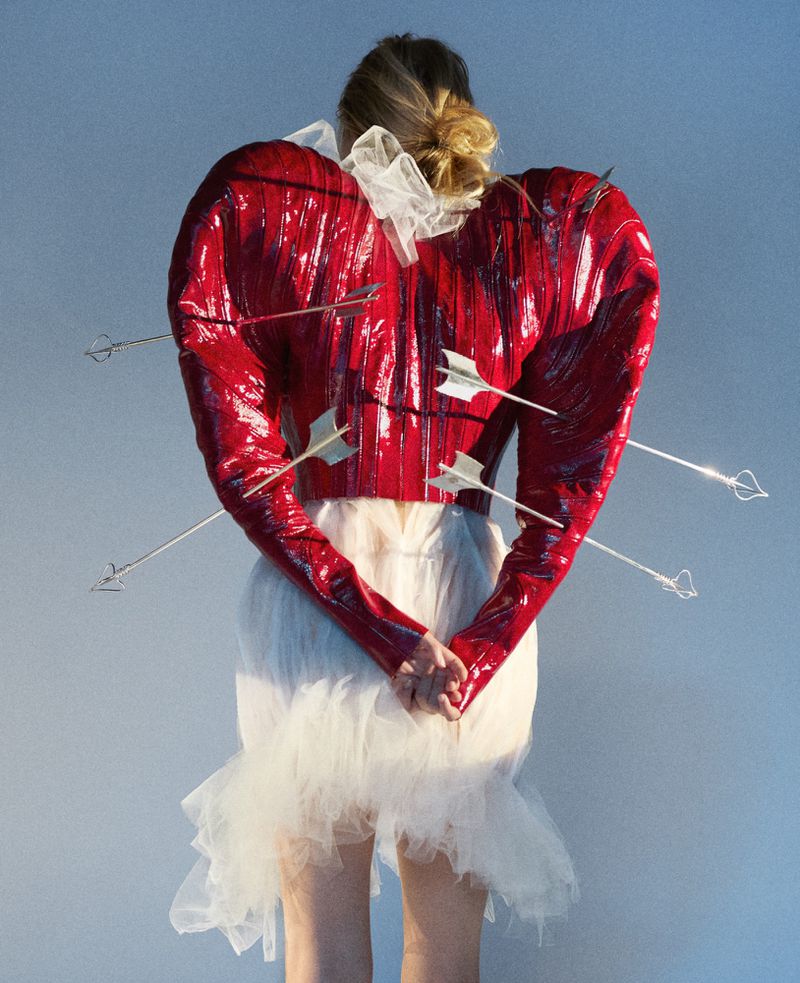 Model seen from behind wearing a glossy red sculptural garment pierced by arrows against a pale grey background