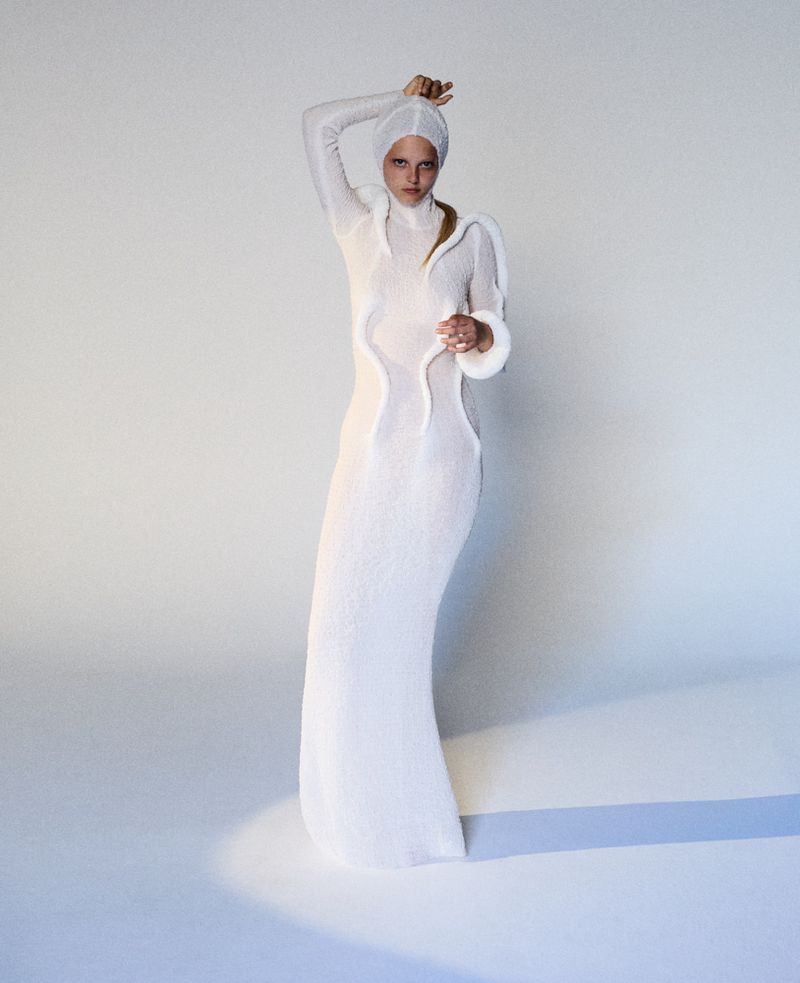 Model wearing a white hooded knit dress with sculptural trim standing against a seamless background for The Unbearable Lightness project