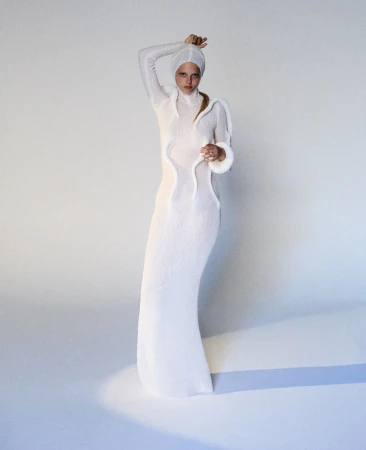 Model wearing a white hooded knit dress with sculptural trim standing against a seamless background for The Unbearable Lightness project