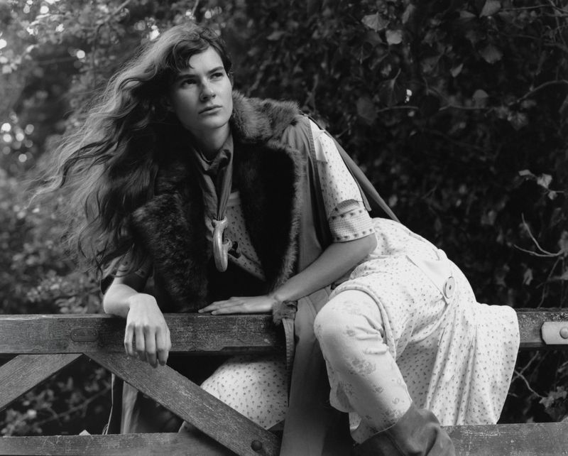 Black and white portrait of a woman leaning on a wooden fence outdoors wearing patterned trousers and a fur-trim vest