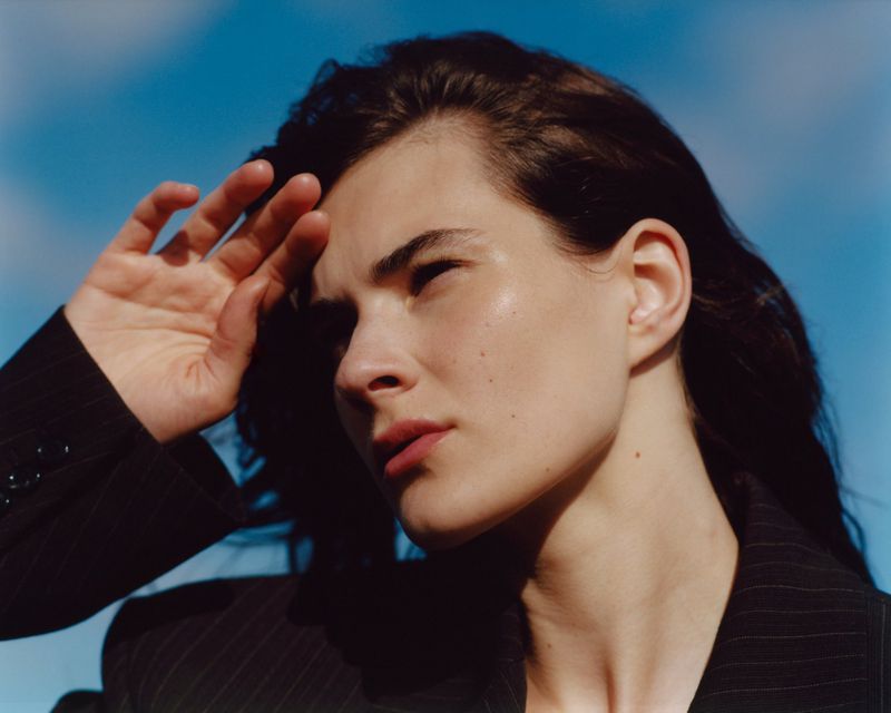 Close-up portrait of a woman shading her eyes against a clear blue sky, wearing a dark blazer.