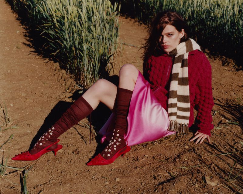 Model seated on dirt ground wearing a red sweater, pink satin skirt, striped scarf, and red kitten heels with floral socks.