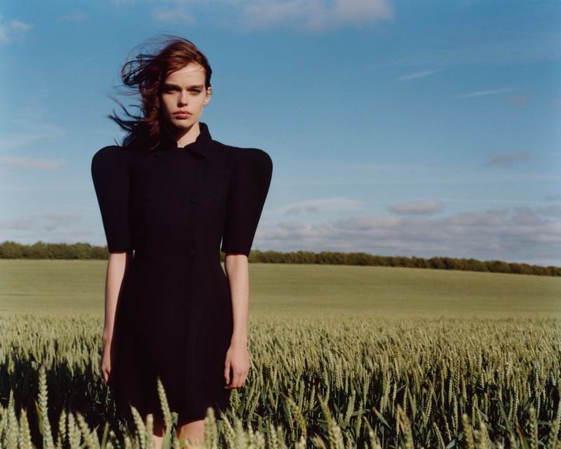 Model in a structured black dress standing in a green wheat field under a blue sky for Heroine Magazine