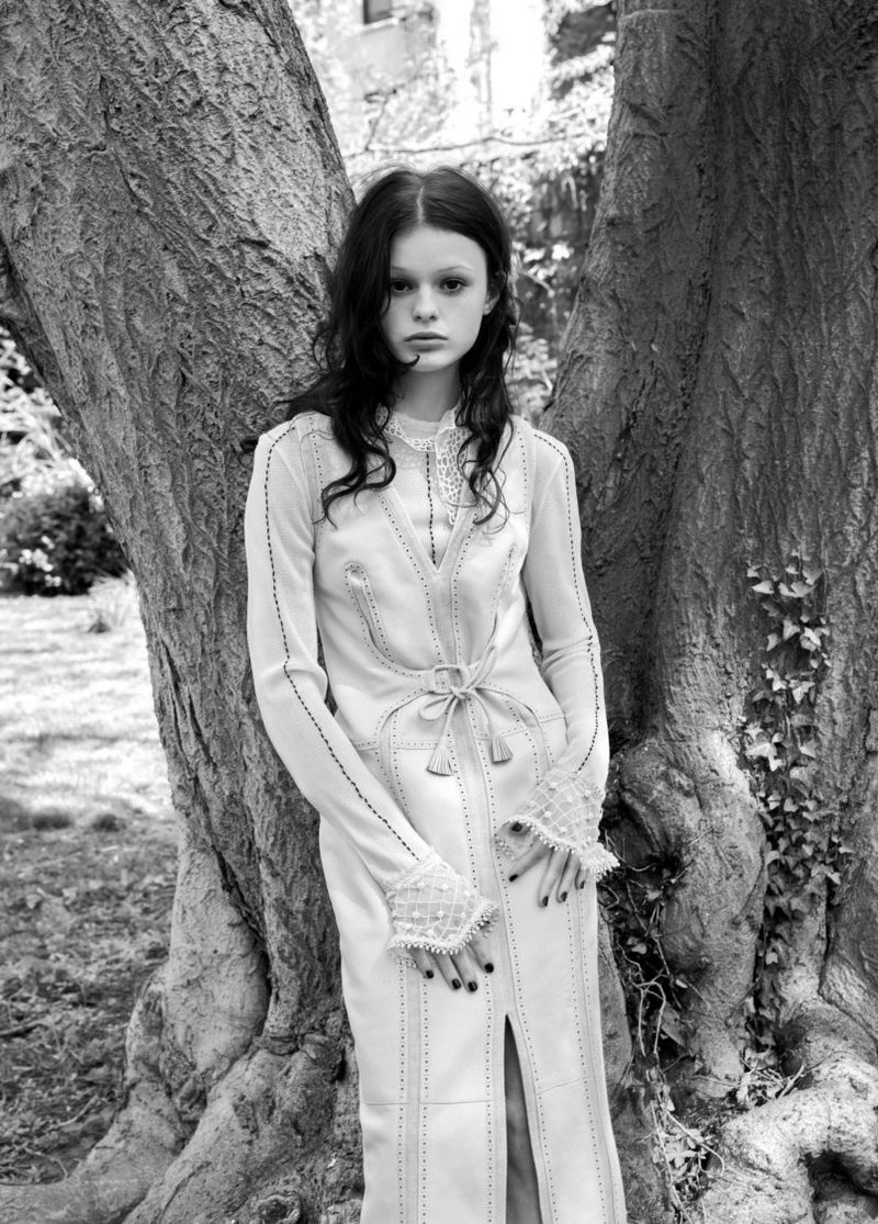 Black and white portrait of a young woman in a long dress, posing in front of a large tree in a natural outdoor setting.
