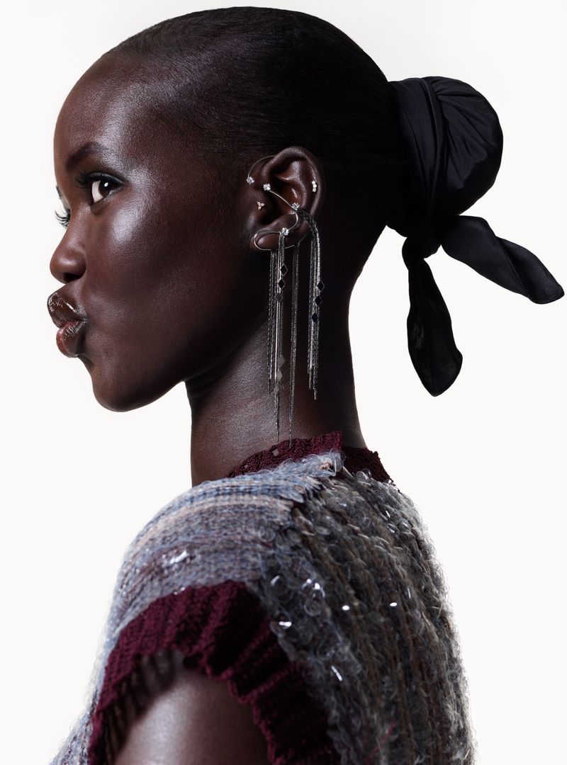 Profile shot of a dark-skinned woman with hair tied in a black scarf, wearing a multi-toned knit top with long silver earrings on a white background.