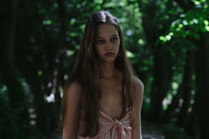 Young female model with long brown hair wearing a pink satin dress standing in a dense green forest