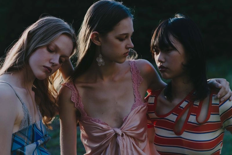 Three female models posing closely together outdoors wearing slip dresses and patterned tops during golden hour.