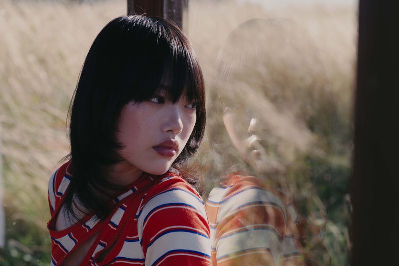 A dark-haired woman in a red and white striped top looking back near a window reflecting a grassy field.