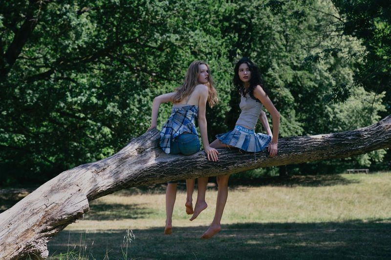 Two young female models sitting on a large fallen tree branch in a park setting