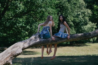Two young female models sitting on a large fallen tree branch in a park setting
