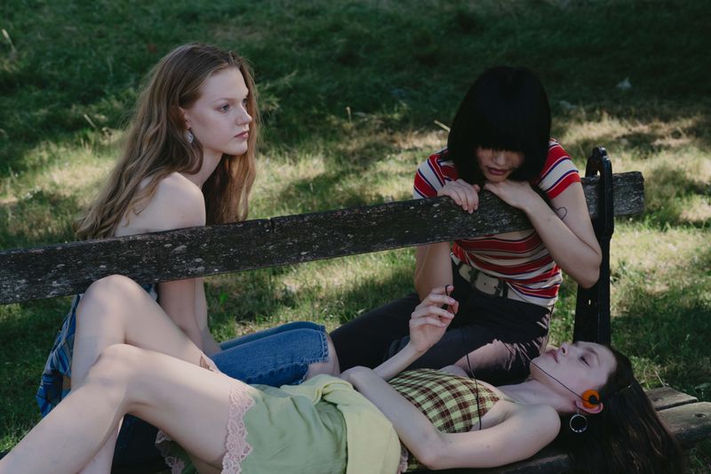 Three young female models relaxing on a wooden park bench in a grassy outdoor setting with natural daylight.
