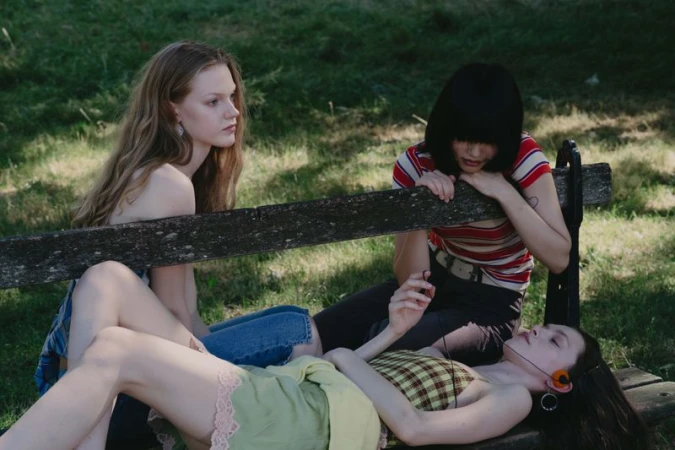 Three young female models relaxing on a wooden park bench in a grassy outdoor setting with natural daylight.
