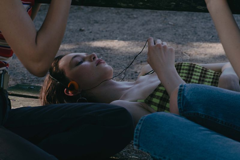 Close-up profile of a young woman lying down wearing vintage headphones, flanked by people in casual clothing outdoors.