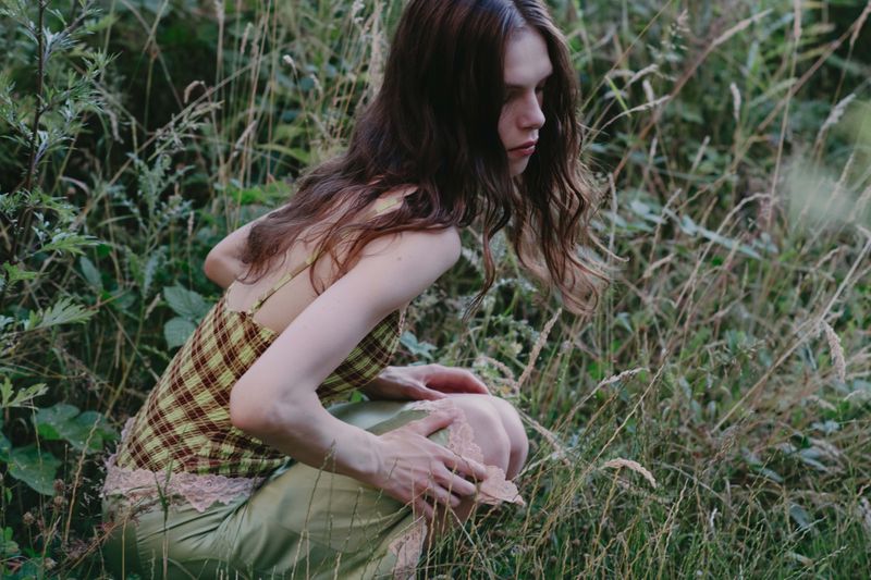 Model crouching in tall grass wearing a plaid camisole and light green skirt for Dreaming Beyond the Fields