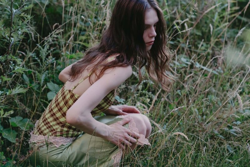 Model crouching in tall grass wearing a plaid camisole and light green skirt for Dreaming Beyond the Fields