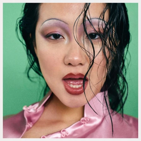 Close-up portrait of an East Asian woman with wet dark hair, purple eyeshadow, thin arched brows, and a pink silk top against green backdrop.