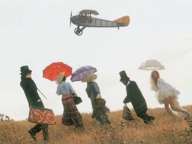 Group of people in period attire running across a grassy field while a vintage biplane flies overhead.