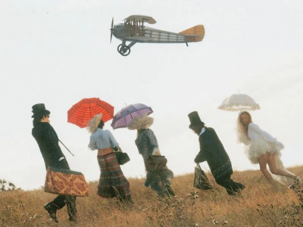 Group of people in period attire running across a grassy field while a vintage biplane flies overhead.