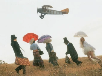 Group of people in period attire running across a grassy field while a vintage biplane flies overhead.