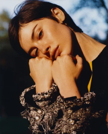 Close-up portrait of a woman resting her chin on her hands, wearing a textured tweed jacket with pearl accents.