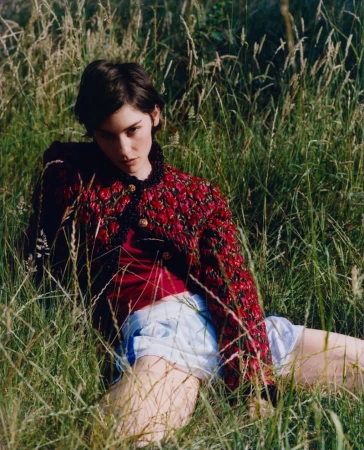 Woman with short dark hair in a red patterned jacket seated outdoors amidst tall wild grass and meadow foliage.