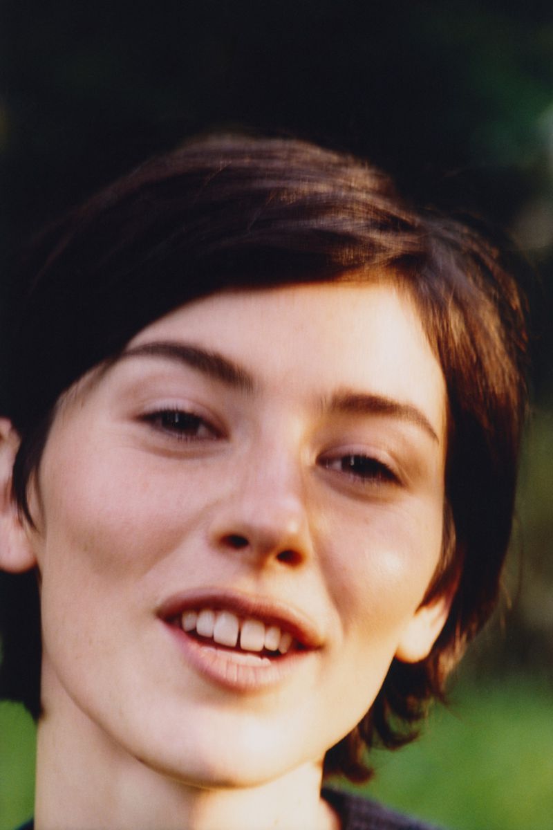 Close-up portrait of a young woman with short dark hair smiling against an outdoor green backdrop