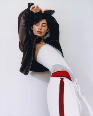 A woman wearing a dark brown leather jacket, white top, and white trousers with red stripe leaning against a white wall.