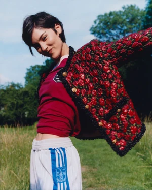 Woman with short dark hair wearing a patterned red jacket and white athletic pants outdoors in a grassy field